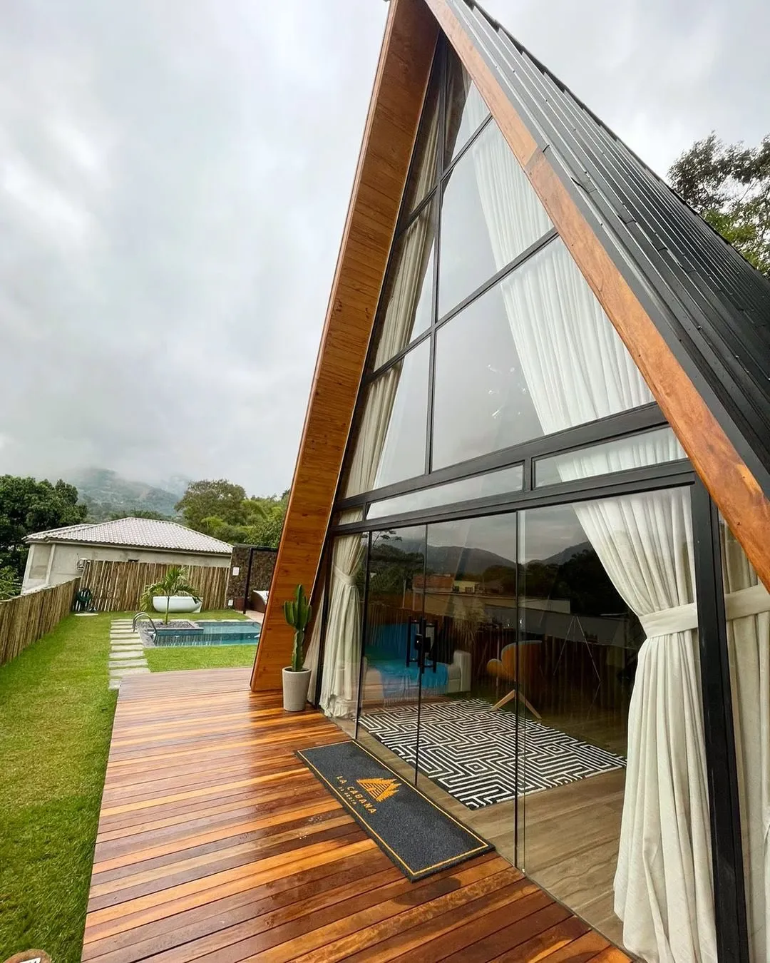 Chalé A-frame pronto, com frente de vidro, cortinas e deck de madeira, ao lado de um gramado com piscina