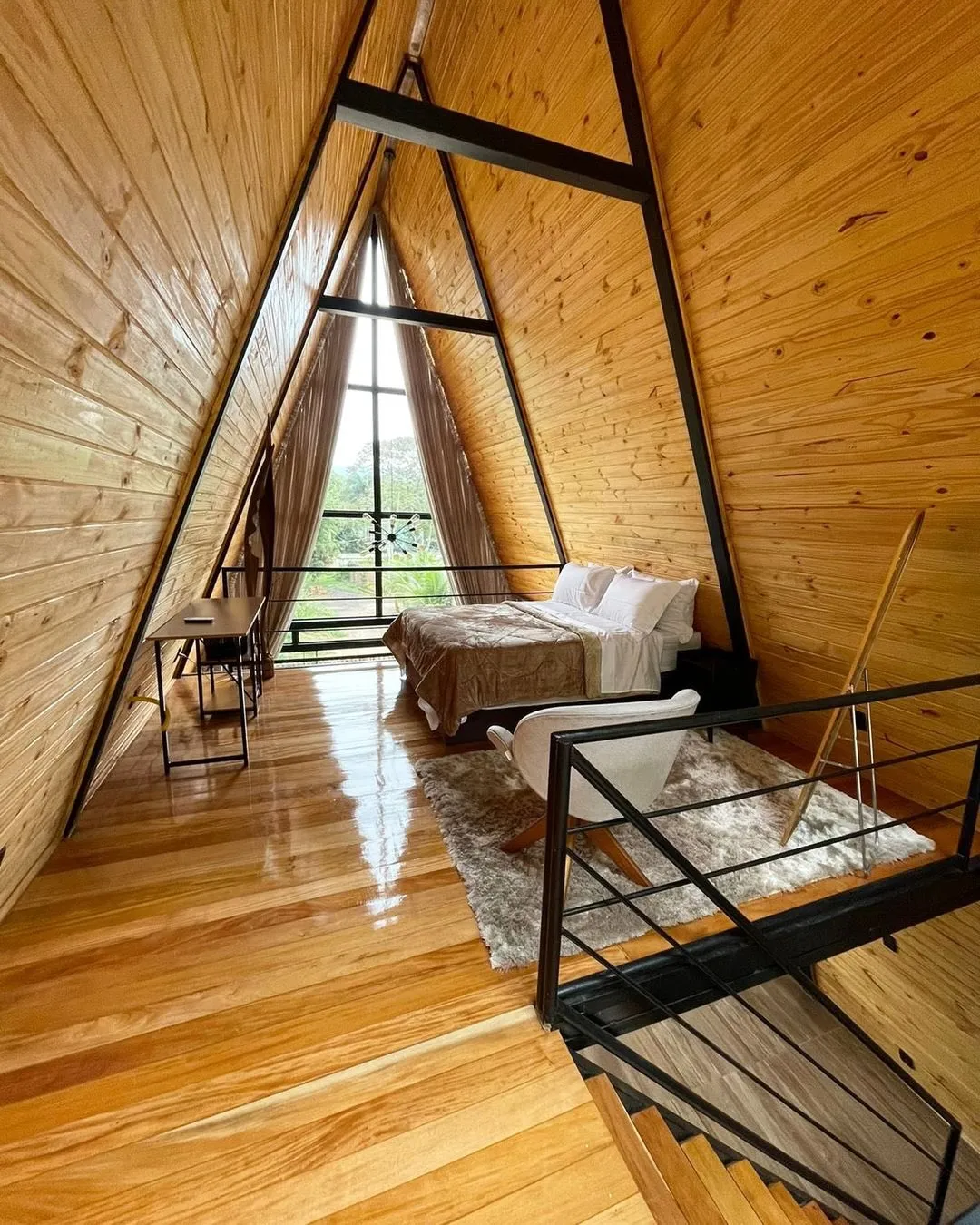 Quarto no mezanino do chalé, sob o telhado em A revestido de madeira, com cama de casal e guarda-corpo preto