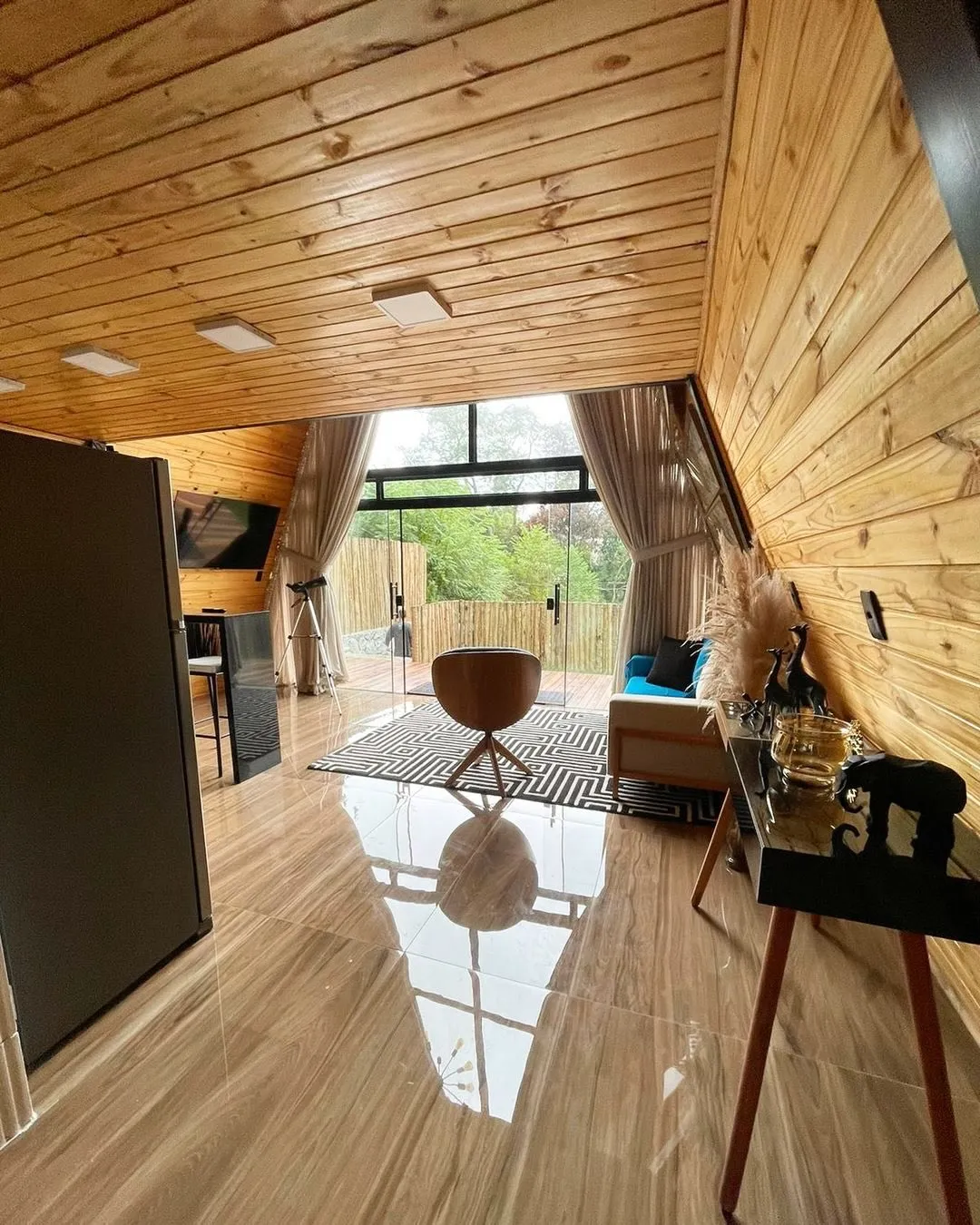 Sala do chalé com forro de madeira, piso claro, sofá e porta de vidro aberta para fora